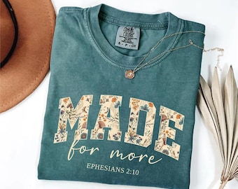 May include: A teal short-sleeved t-shirt with a crew neckline. The front features the word MADE in large block letters filled with a floral pattern, followed by the text for more and EPHESIANS 2:10 in a light-colored script and block font.