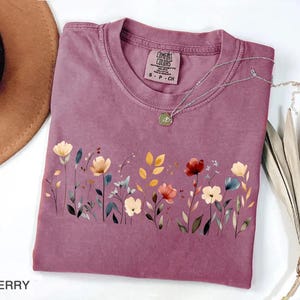 Peut inclure: T-shirt de couleur baie avec un motif floral sur le devant. Le t-shirt a un col rond et des manches courtes. Un collier argent&eacute; avec un pendentif repose sur le t-shirt. Le t-shirt est &eacute;tiquet&eacute; "Comfort Colors".