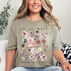 May include: A khaki-coloured short-sleeve t-shirt featuring a central graphic of various pink, purple, and yellow flowers, green foliage, and butterflies. The text "IT IS WELL WITH MY SOUL" is printed in black capital letters above and below the floral design.