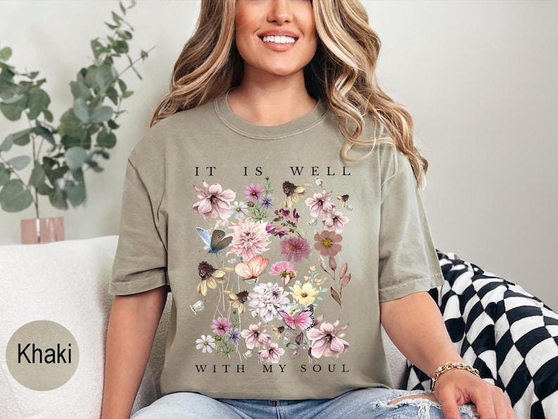 May include: A khaki-coloured short-sleeve t-shirt featuring a central graphic of various pink, purple, and yellow flowers, green foliage, and butterflies. The text "IT IS WELL WITH MY SOUL" is printed in black capital letters above and below the floral design.