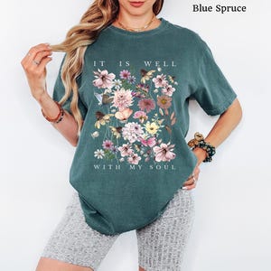 May include: A blue spruce coloured short-sleeve t-shirt featuring a graphic print of various colourful flowers and butterflies. The text "IT IS WELL WITH MY SOUL" is printed in white capital letters above and below the floral arrangement.