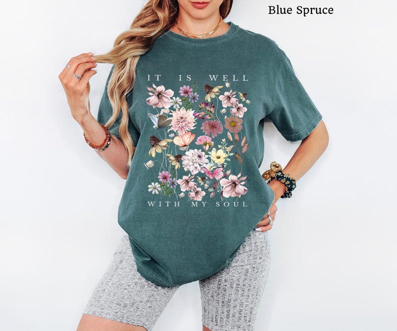 May include: A blue spruce coloured short-sleeve t-shirt featuring a graphic print of various colourful flowers and butterflies. The text "IT IS WELL WITH MY SOUL" is printed in white capital letters above and below the floral arrangement.