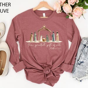 K&ouml;nnte beinhalten: Lang&auml;rmeliges Shirt in Heather Mauve mit einer Aquarell-Weihnachtskrippe und dem Text "the greatest gift of all" und Matth&auml;us 1:21. Das Shirt hat einen Rundhalsausschnitt und einen geknoteten Saum. Die Worte "Heather Mauve" sind oben links aufgedruckt.