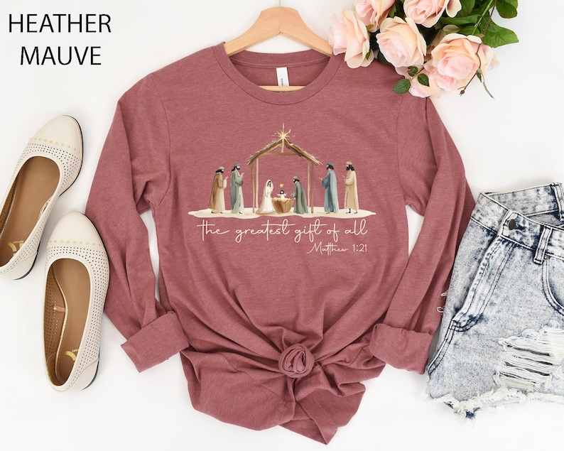 K&ouml;nnte beinhalten: Lang&auml;rmeliges Shirt in Heather Mauve mit einer Aquarell-Weihnachtskrippe und dem Text "the greatest gift of all" und Matth&auml;us 1:21. Das Shirt hat einen Rundhalsausschnitt und einen geknoteten Saum. Die Worte "Heather Mauve" sind oben links aufgedruckt.