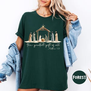 K&ouml;nnte beinhalten: Forstgr&uuml;nes T-Shirt mit einer Aquarellillustration einer Krippenszene und dem Text "the greatest gift of all" und "Matthew 1:21". Das Design umfasst Figuren und einen Stall. Das Shirt ist ein bequemer, l&auml;ssiger Stil.