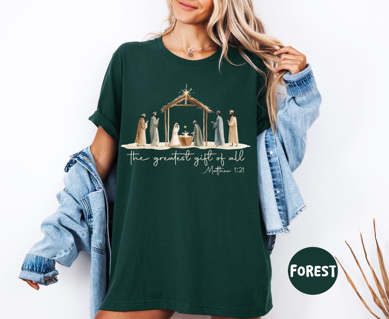 K&ouml;nnte beinhalten: Forstgr&uuml;nes T-Shirt mit einer Aquarellillustration einer Krippenszene und dem Text "the greatest gift of all" und "Matthew 1:21". Das Design umfasst Figuren und einen Stall. Das Shirt ist ein bequemer, l&auml;ssiger Stil.