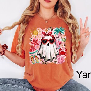 Peut inclure: Un t-shirt orange br&ucirc;l&eacute; avec un motif central repr&eacute;sentant un fant&ocirc;me blanc portant un bandeau rouge et des lunettes de soleil en forme de c&oelig;ur. Le design inclut des &eacute;l&eacute;ments estivaux comme un palmier, une &eacute;toile de mer et un crabe.