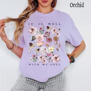 May include: An orchid-coloured short-sleeve t-shirt featuring a central graphic of various flowers and butterflies in shades of pink, yellow, and blue. The text "IT IS WELL" is printed above the floral design, and "WITH MY SOUL" is printed below it.