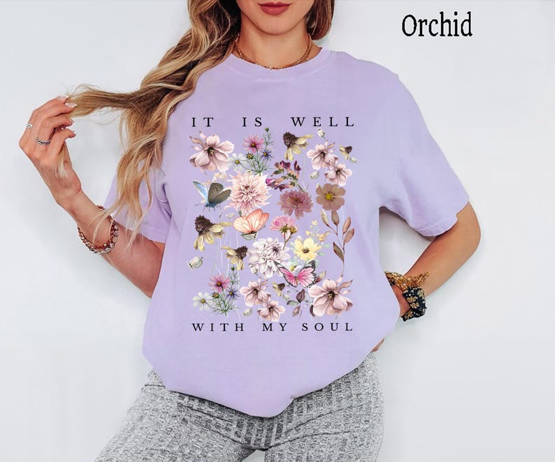 May include: An orchid-coloured short-sleeve t-shirt featuring a central graphic of various flowers and butterflies in shades of pink, yellow, and blue. The text "IT IS WELL" is printed above the floral design, and "WITH MY SOUL" is printed below it.