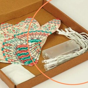 K&ouml;nnte beinhalten: Ein brauner Pappkarton enth&auml;lt ein Set dekorativer Papierf&auml;cher-Lichter. Die F&auml;cher haben ein Blumendesign in Rot, T&uuml;rkis und Rosa. Ein Akku und Lichterketten befinden sich ebenfalls in der Box.