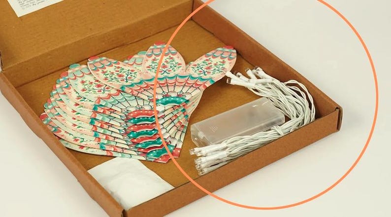 K&ouml;nnte beinhalten: Ein brauner Pappkarton enth&auml;lt ein Set dekorativer Papierf&auml;cher-Lichter. Die F&auml;cher haben ein Blumendesign in Rot, T&uuml;rkis und Rosa. Ein Akku und Lichterketten befinden sich ebenfalls in der Box.