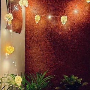 K&ouml;nnte beinhalten: Eine Lichterkette mit gelb get&ouml;nten, dekorativen Laternen. Die Lichter sind vor einer strukturierten, rotbraunen Wand angebracht. Gr&uuml;ne Pflanzen im Vordergrund schaffen eine warme und einladende Atmosph&auml;re.