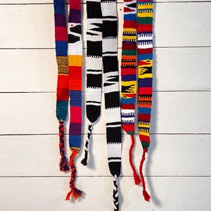 May include: Six colorful woven belts with different patterns, including stripes, geometric designs, and a black and white checkerboard pattern. The belts are hanging against a white wooden background.