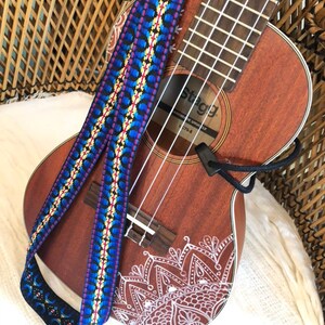May include: A brown ukulele with a patterned blue, yellow, and red strap. The ukulele has a white mandala design on the body. The strap is woven with a geometric design. The ukulele is resting on a white surface.