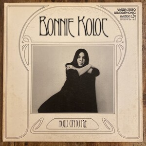 May include: A black and white photo of a woman with long dark hair, wearing a black dress, with her arms crossed. The photo is on a white album cover with the text "Bonnie Koloc" and "Hold On To Me" in black.