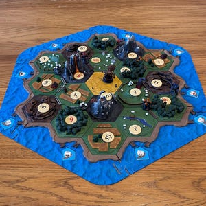 Könnte beinhalten: Ein Brettspiel mit sechseckigem Layout, das Geländefelder mit Feldern, Wäldern, Bergen und Wasser zeigt. Das Spielbrett ist von einem blauen Rand umgeben, und die Geländefelder haben Zahlen und kleine Strukturen.