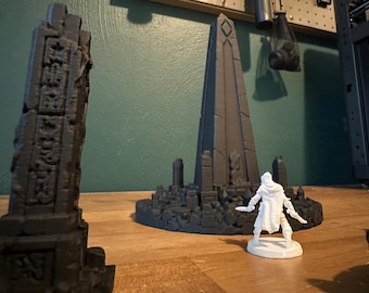 May include: A dark grey 3D printed obelisk with intricate carved symbols stands in the foreground, while a larger dark grey obelisk structure sits in the background with a small white hooded figurine standing in front of it on a wooden surface.