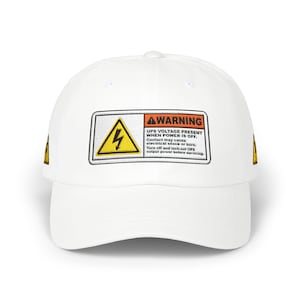 May include: White baseball cap with a curved peak. The cap has a rectangular patch with a yellow lightning bolt symbol and the text "WARNING UPS VOLTAGE PRESENT WHEN POWER IS OFF." A small yellow triangle is on the side.