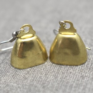 May include: A pair of gold-coloured bell-shaped earrings featuring a smooth metallic finish and a small loop at the top. Each bell is attached to a silver-toned metal hook wire, designed for hanging from the earlobe with a simple, functional structure.