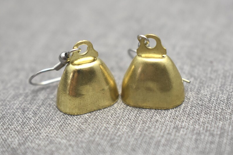 May include: A pair of gold-coloured bell-shaped earrings featuring a smooth metallic finish and a small loop at the top. Each bell is attached to a silver-toned metal hook wire, designed for hanging from the earlobe with a simple, functional structure.