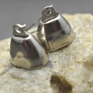 May include: Two small, polished silver-toned metal bells with a smooth, rounded, bell-shaped body and a small loop at the top for attachment. The bells have a reflective, metallic surface and are positioned together, showing their curved form.