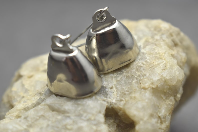 May include: Two small, polished silver-toned metal bells with a smooth, rounded, bell-shaped body and a small loop at the top for attachment. The bells have a reflective, metallic surface and are positioned together, showing their curved form.