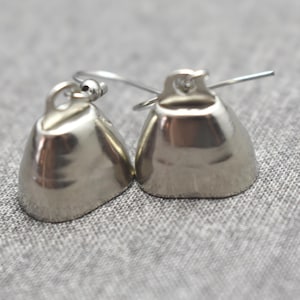 May include: A pair of silver-toned bell-shaped earrings featuring a smooth, reflective metallic finish. Each earring is equipped with a thin, curved wire hook for attachment, showcasing a simple, rounded design with a small loop at the top.