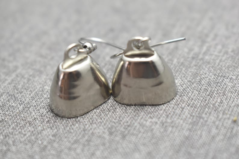 May include: A pair of silver-toned bell-shaped earrings featuring a smooth, reflective metallic finish. Each earring is equipped with a thin, curved wire hook for attachment, showcasing a simple, rounded design with a small loop at the top.