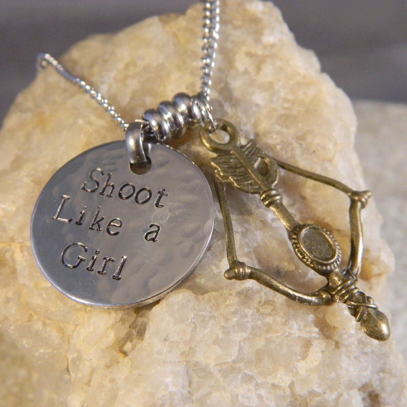 May include: A silver-toned metal necklace featuring a round, hammered pendant stamped with the text Shoot Like a Girl and a separate bronze-toned charm shaped like a bow and arrow, both suspended from a silver-toned ball chain necklace.