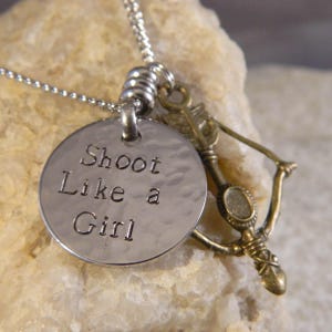 May include: A silver-toned metal necklace featuring a round, hammered pendant stamped with the text Shoot Like a Girl, accompanied by a small, antique brass-colored charm shaped like a bow and arrow, all suspended from a thin silver chain.