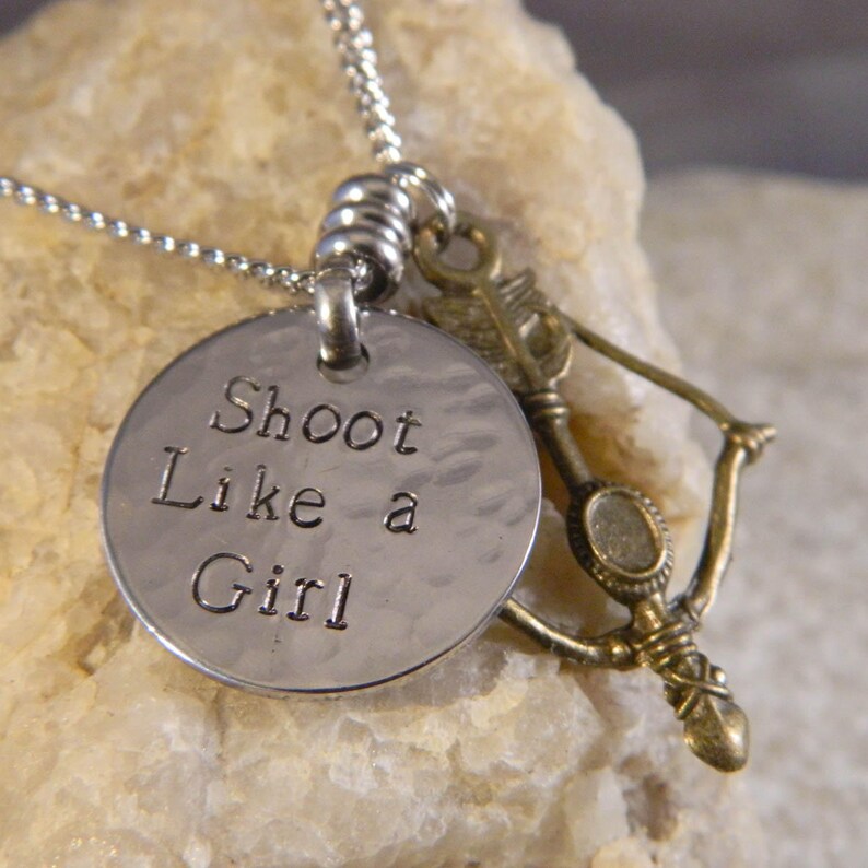 May include: A silver-toned metal necklace featuring a round, hammered pendant stamped with the text Shoot Like a Girl, accompanied by a small, antique brass-colored charm shaped like a bow and arrow, all suspended from a thin silver chain.