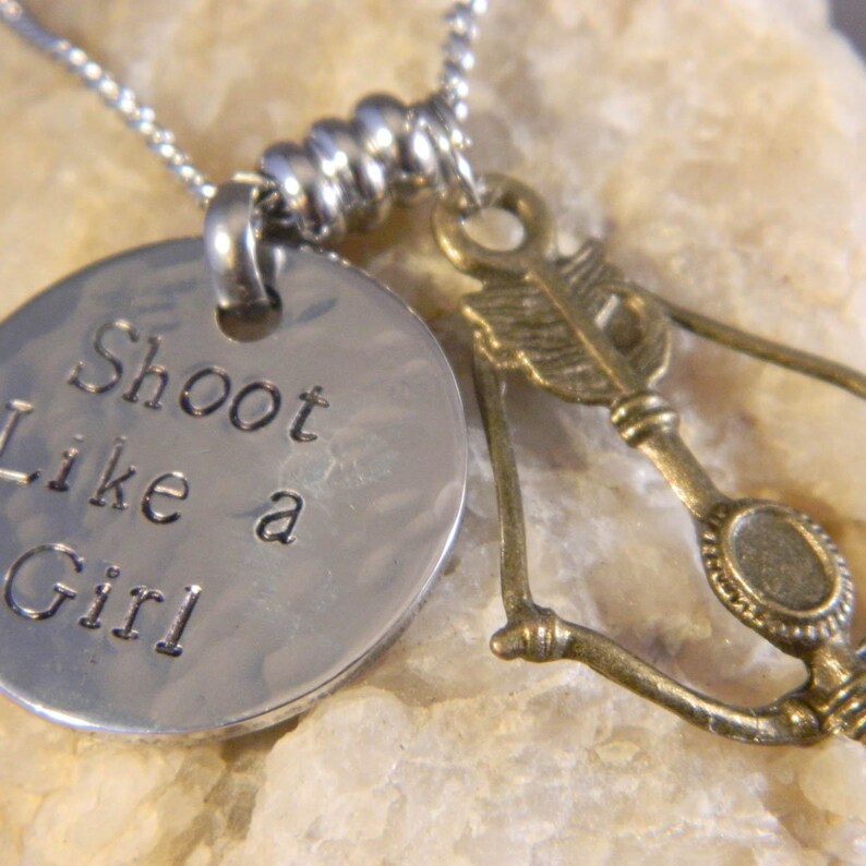 May include: A silver-toned metal necklace featuring a round, hammered pendant stamped with the text Shoot Like a Girl. Beside the pendant hangs a bronze-toned charm shaped like a bow and arrow. The charms are suspended from a silver-toned chain.