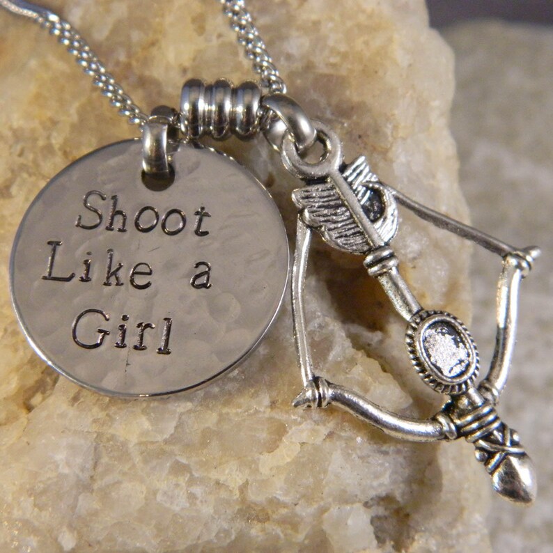 May include: A silver-toned necklace featuring a round, hammered metal pendant stamped with the text "Shoot Like a Girl" and a separate charm shaped like a bow and arrow. The jewelry is suspended from a delicate silver-toned chain link necklace.