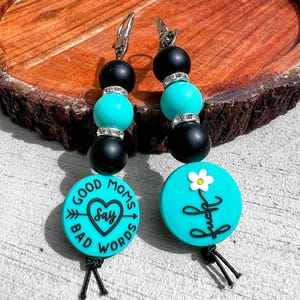 May include: A pair of beaded earrings with black and turquoise beads, and silver-colored metal findings. One earring has a turquoise disc with the words "GOOD MOMS SAY BAD WORDS" and the other has a turquoise disc with a white flower and the word "fuck".