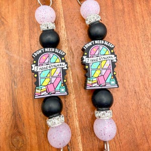 May include: Two beaded lanyards with silver-tone clips. Each lanyard features a pink speckled bead, a silver rhinestone spacer, a black bead, and a charm with the text "I don't need sleep, I need answers" above a colorful book and window design.