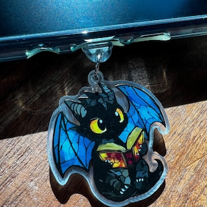 May include: A clear acrylic charm featuring a black dragon with blue wings and yellow eyes, reading a book. The dragon is holding a book with red and yellow pages. The charm has a silver-colored metal loop for attachment.