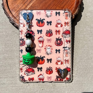 May include: A clear phone case with a pink Halloween-themed pattern featuring pumpkins, bows, ghosts, and coffee cups. A charm with a green witch's cauldron, black and white beads, and a blue heart is attached.
