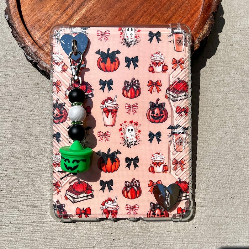 May include: A clear phone case with a pink Halloween-themed pattern featuring pumpkins, bows, ghosts, and coffee cups. A charm with a green witch's cauldron, black and white beads, and a blue heart is attached.