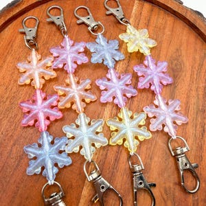 May include: Four keychains featuring translucent, glittery snowflake charms in pastel shades of pink, blue, yellow, and orange. Each keychain is constructed with a silver-toned metal lobster clasp at both ends, connecting a vertical row of three snowflakes.