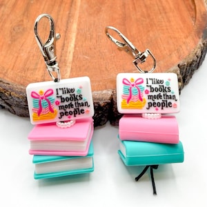May include: Two keychains with a book theme. Each keychain has a white rectangular top with the text "I like books more than people" and a stack of colorful books in pink, blue, and white. The keychains have silver clasps.