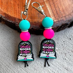 May include: Two keychains with silver clasps. Each keychain has a teal bead, a pink bead, and a black charm with the text "Smiette Books & Coffee Please" in white and pink. The charms are shaped like a tombstone with an open book.