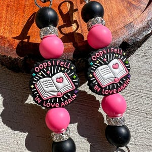 May include: Two beaded lanyards with black, pink, and silver accents. Each lanyard features a charm with the text "Oops I Fell In Love Again" and an open book graphic with a heart. The lanyards have silver-colored clips.