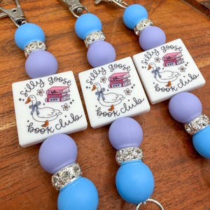 May include: Three keychains featuring a square white charm with a printed illustration of a goose wearing a blue ribbon next to a stack of books and the text silly goose book club. The charms are accented by light blue and lavender beads.