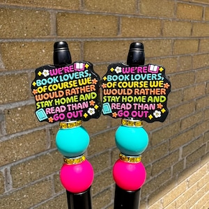 May include: Two black pens with decorative toppers. Each topper has a black, cloud-shaped design with the text "WE'RE BOOK LOVERS, OF COURSE WE WOULD RATHER STAY HOME AND READ THAN GO OUT." The pens have turquoise and pink beads.