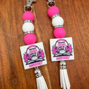 May include: Two keychains with pink and white beads, silver accents, and white tassels. Each keychain has a square charm with the text "WHO NEEDS FRIENDS WHEN YOU HAVE NARRATORS?" and a floral and headphone design.