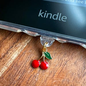 May include: A red cherry charm with a green leaf hangs from a clear Kindle case. The word "kindle" is visible on the screen. The charm is attached with a gold-colored clasp. The background is a wooden surface.