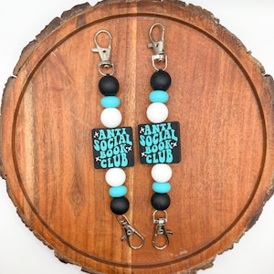 May include: Two beaded lanyards with silver-tone clasps. Each lanyard features black, turquoise, and white beads, with a black square pendant that reads "ANTI SOCIAL BOOK CLUB" in turquoise text.