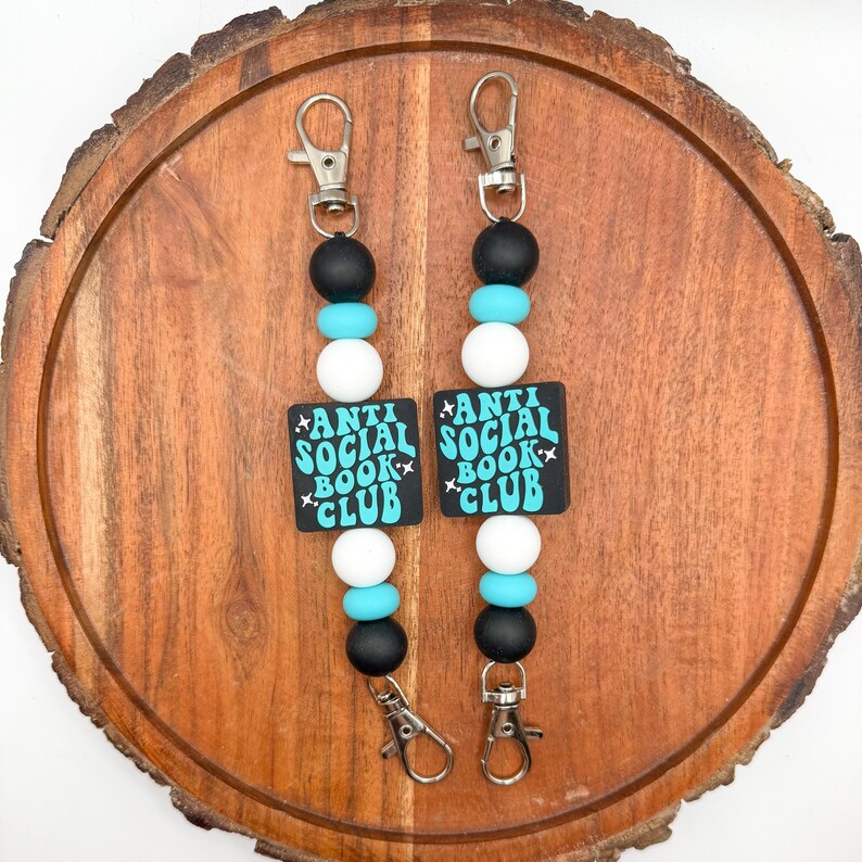 May include: Two beaded lanyards with silver-tone clasps. Each lanyard features black, turquoise, and white beads, with a black square pendant that reads "ANTI SOCIAL BOOK CLUB" in turquoise text.