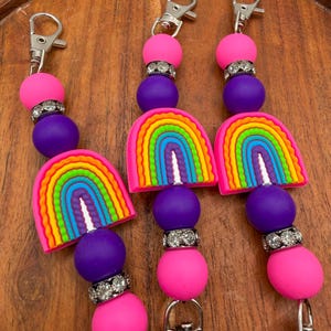 May include: Three keychains with a rainbow theme. Each keychain features a silver clasp, pink and purple beads, a rhinestone spacer, and a colorful rainbow charm. The keychains are displayed on a wooden surface.