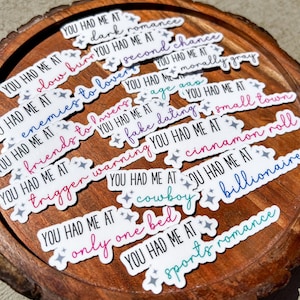 May include: A collection of white stickers with phrases like "You Had Me At Dark Romance" and "You Had Me At Enemies to Lovers." The stickers have colorful text and star accents, arranged on a wooden surface.